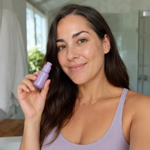 A smiling woman holding up a small lilac skincare bottle in a bright bathroom.