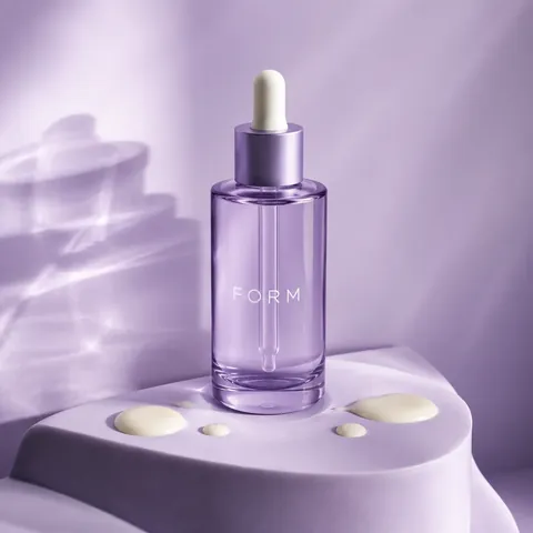 A lilac skincare bottle standing on a soft purple plinth in gentle window light.